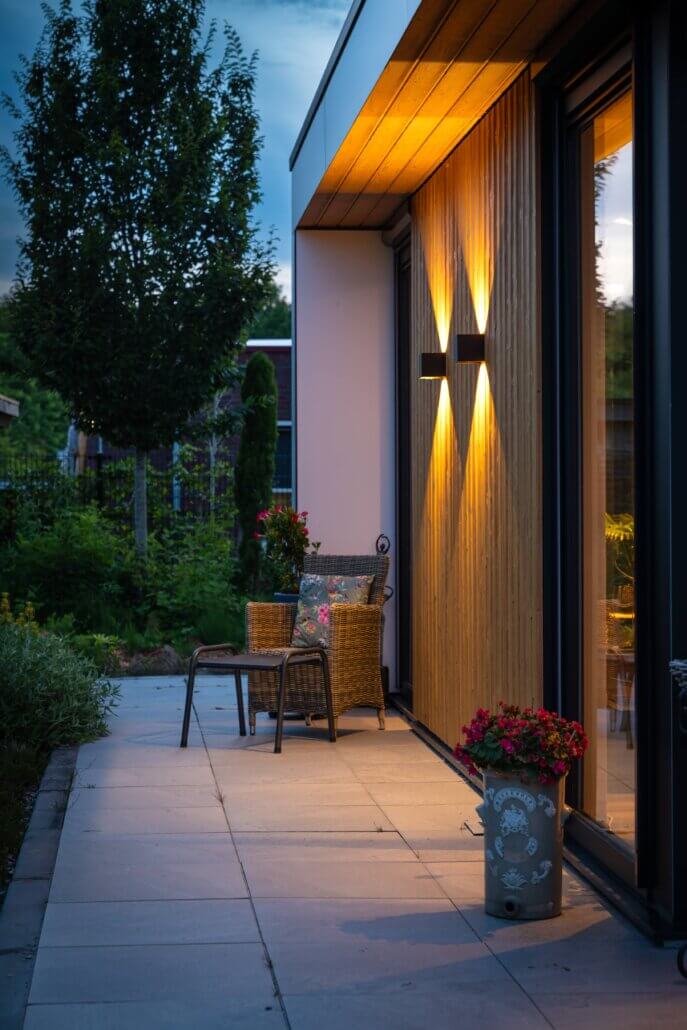 Moderne mantelzorgwoning van Solilux in Lelystad in avondlicht, met L-vormige opzet, gevelbekleding in warme houttinten, bakstenen accentwand, zwarte pergolaconstructie en sfeervolle verlichting.