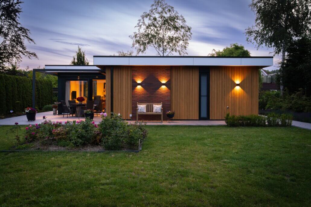 Moderne mantelzorgwoning van Solilux in Lelystad in avondlicht, met L-vormige opzet, gevelbekleding in warme houttinten, bakstenen accentwand, zwarte pergolaconstructie en sfeervolle verlichting.